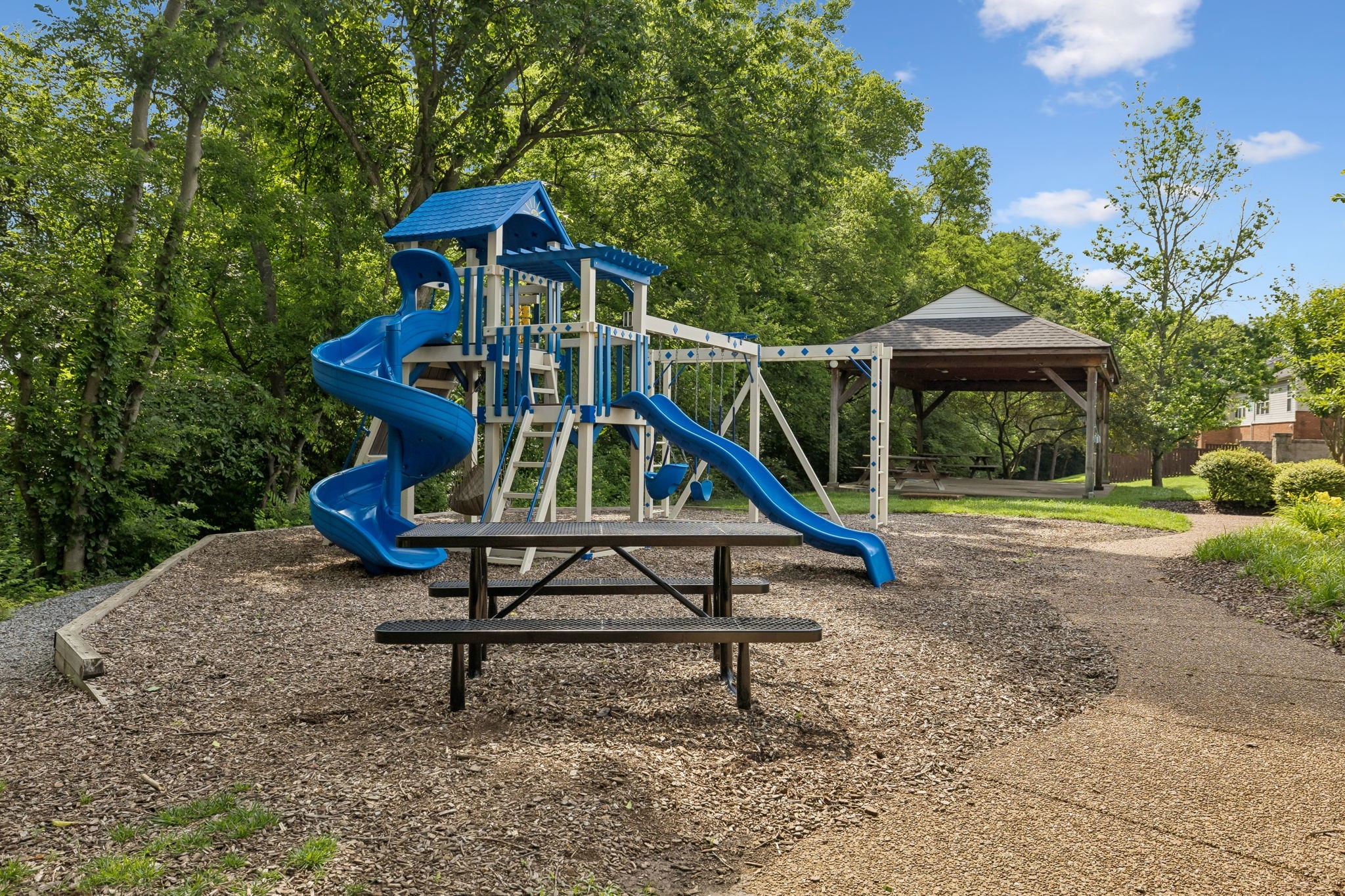 Outdoor playground with blue slides and swings, a picnic table, and a covered pavilion among trees.