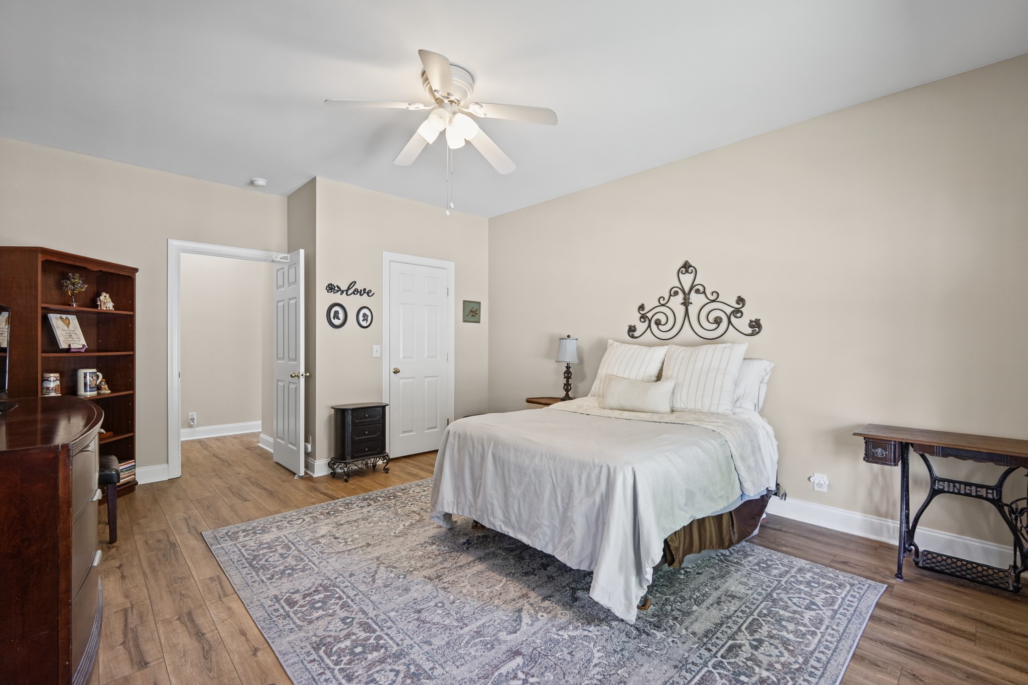 Furnished bedroom with a white bed, ornate metal wall decor, ceiling fan, and patterned rug.
