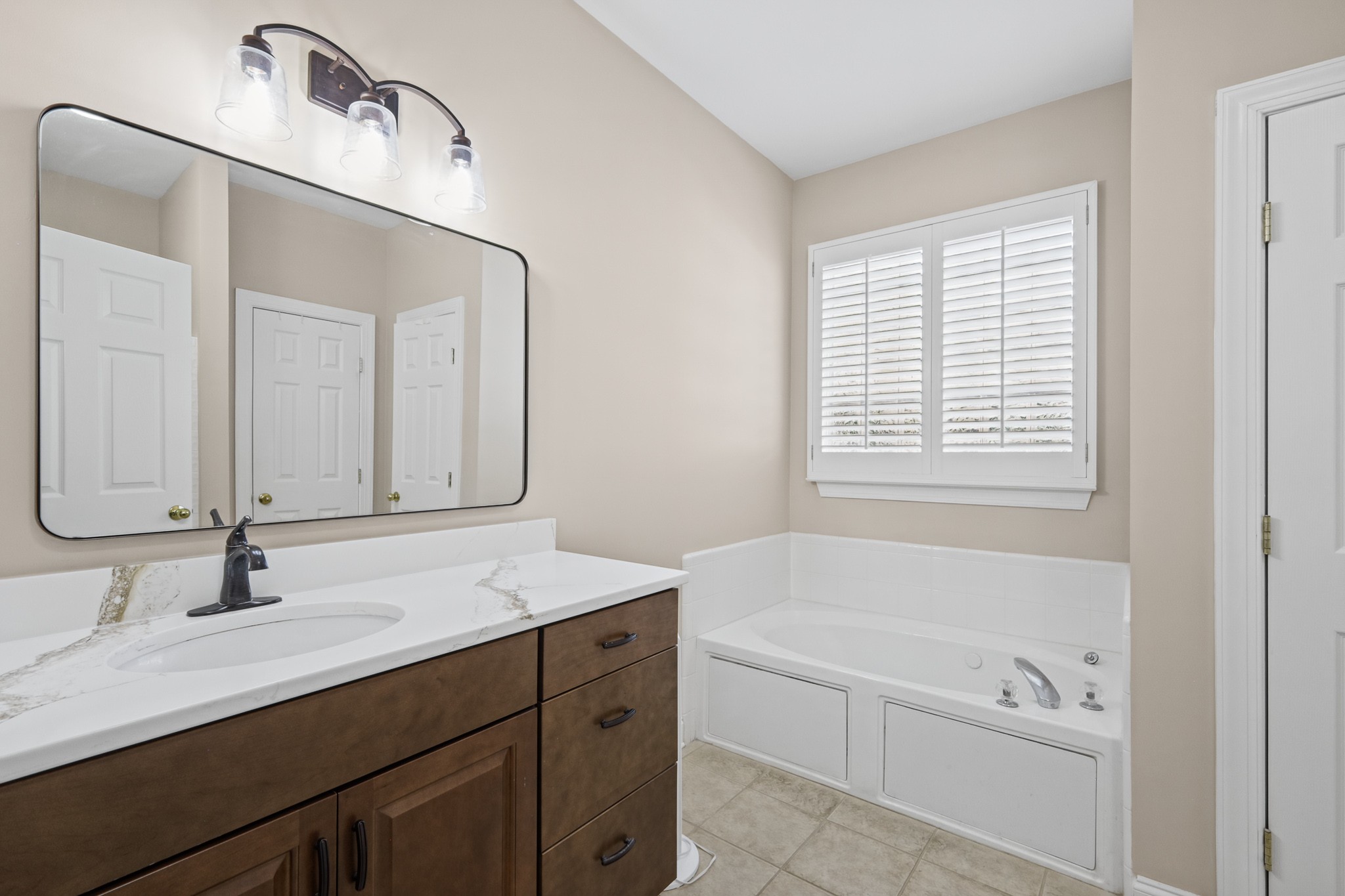 Bathroom with a dark wood vanity, marble-look countertop, and built-in soaking tub beneath a shuttered window.