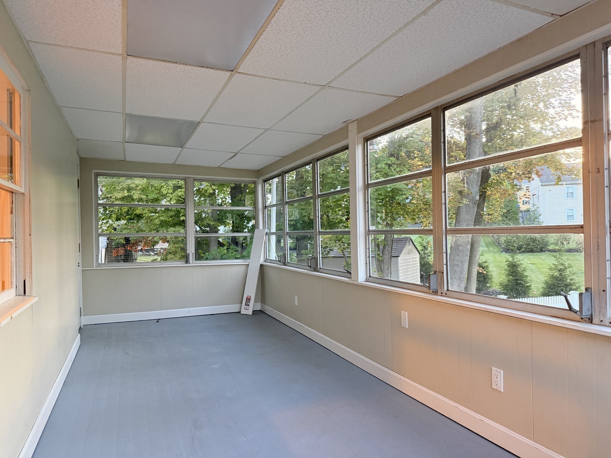 Empty enclosed sunroom with gray flooring and large windows overlooking a leafy yard.