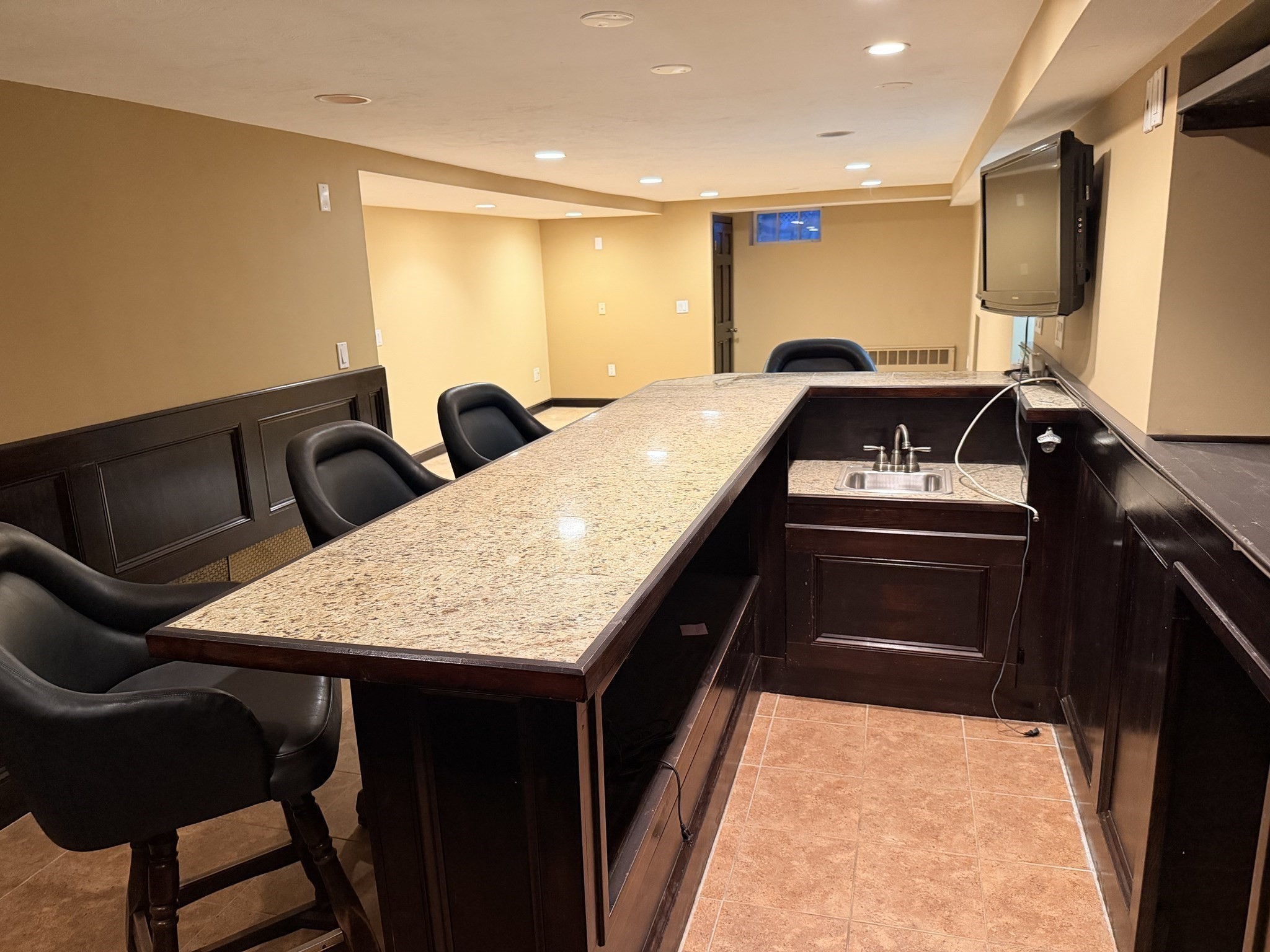Basement bar with granite counter, dark cabinetry, black stools, sink, and wall-mounted television.