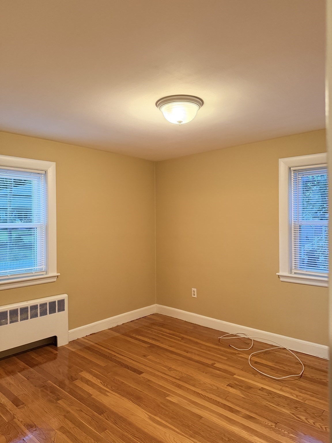 Empty room with hardwood floors, yellow walls, two blinded windows, and a wall-mounted radiator.