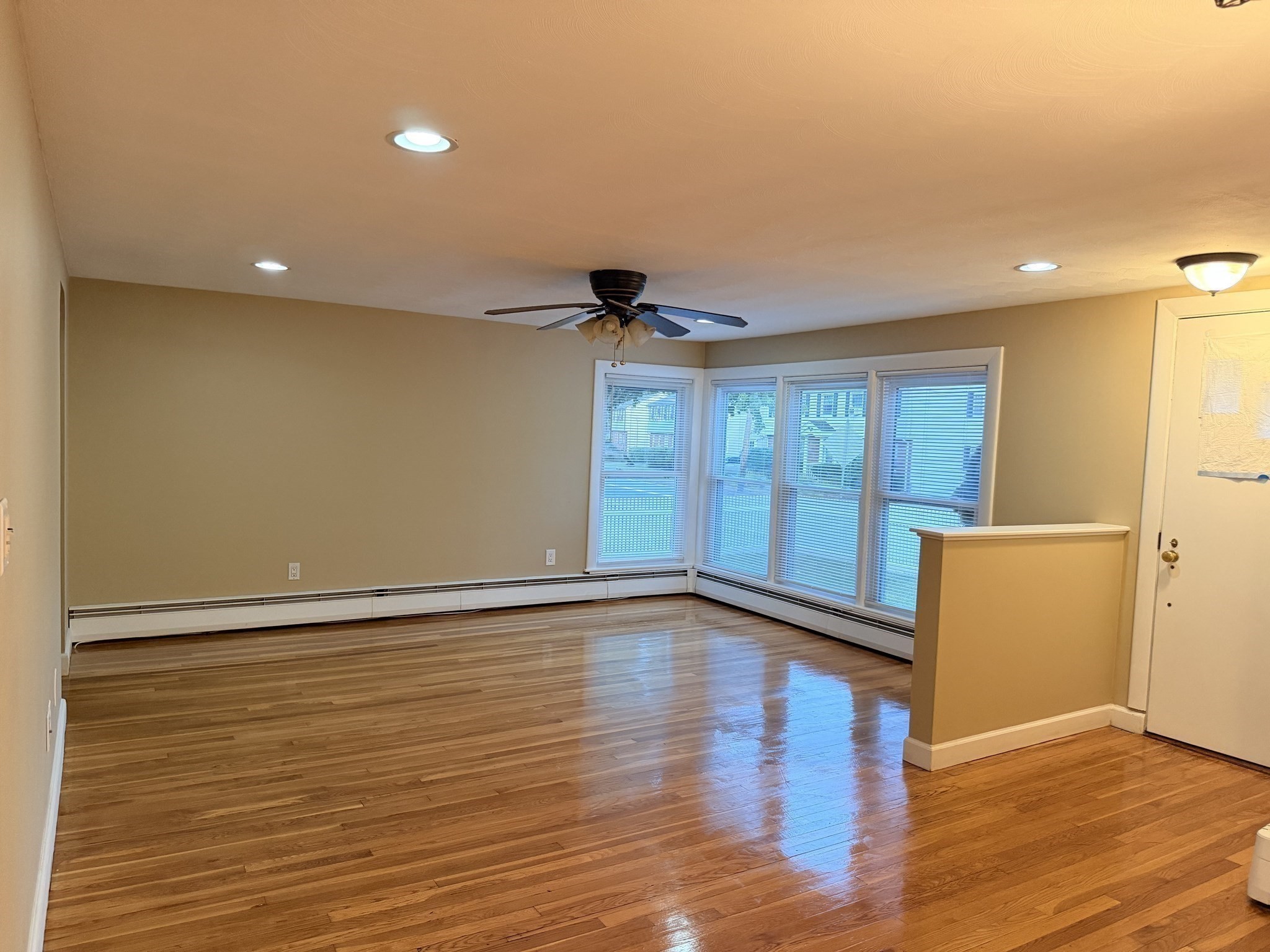 Vacant living room with glossy hardwood floors, corner windows with blinds, and a ceiling fan.