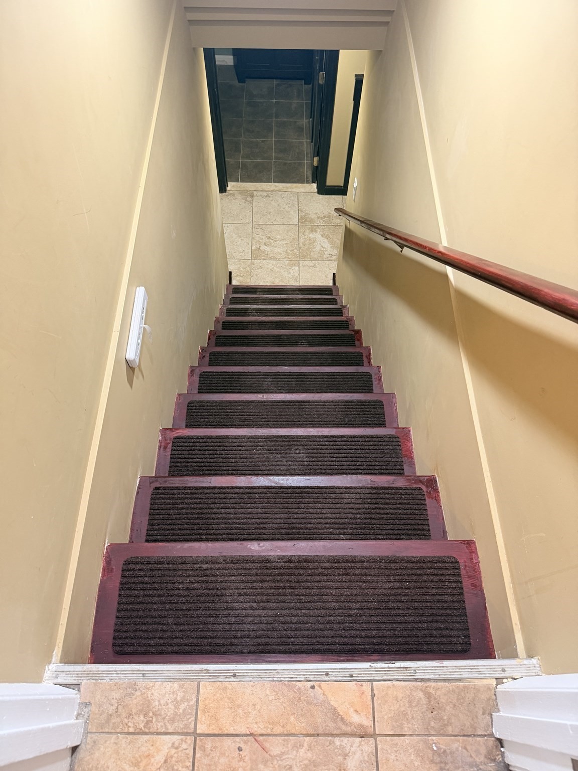 Narrow staircase with dark carpet runners and a wooden handrail leading to a tiled landing.