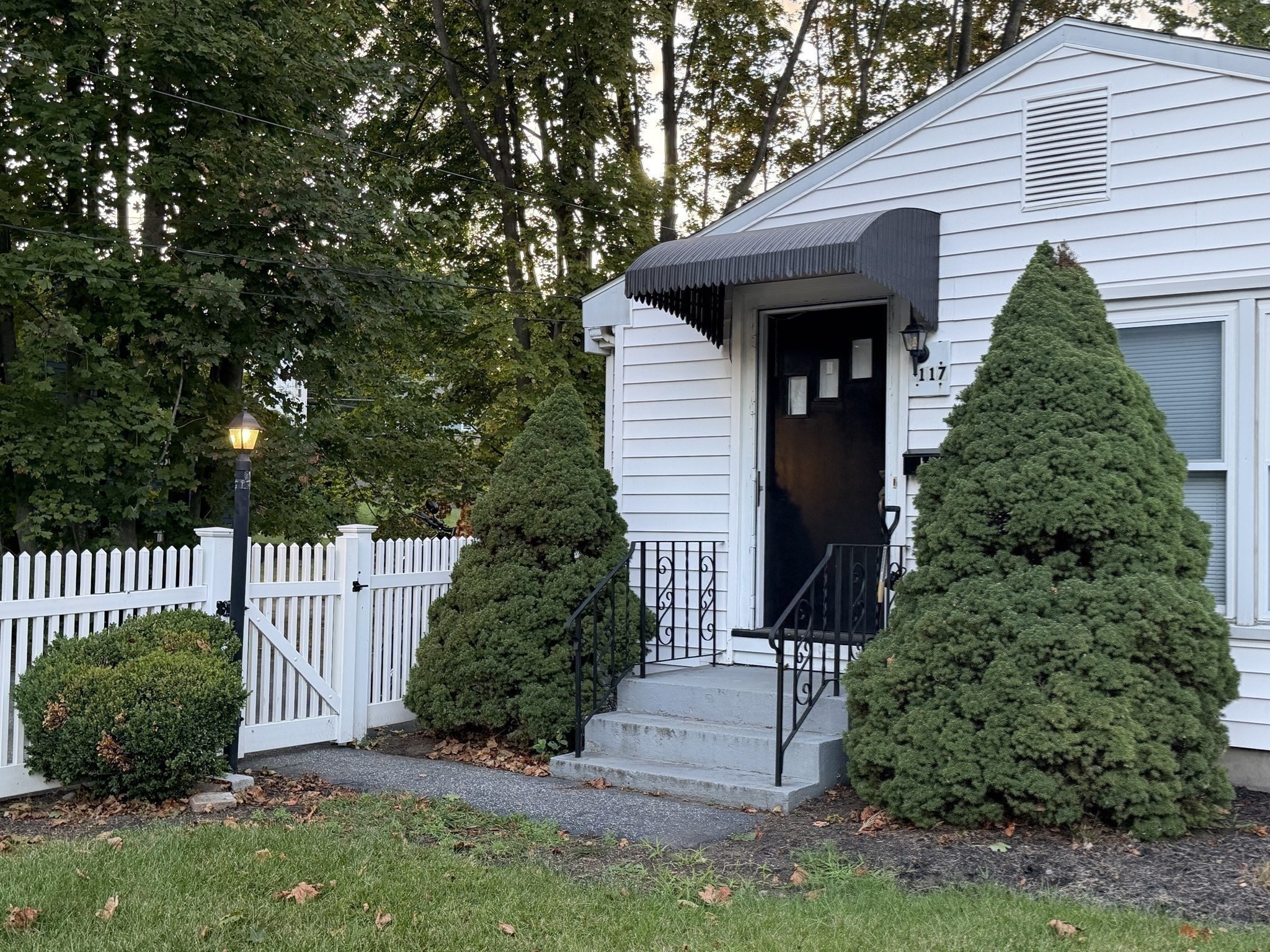 White-sided building entrance with a black awning, concrete steps, picket fence, and evergreen shrubs.