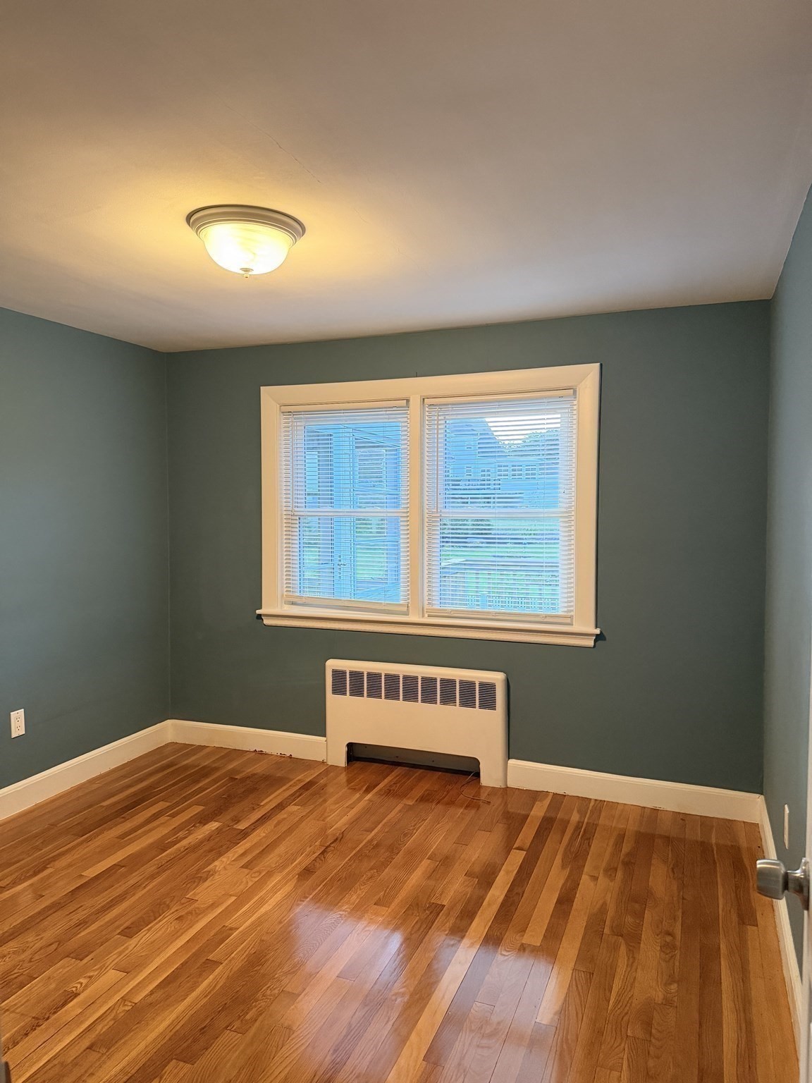Empty room with teal walls, hardwood floors, and a double window above a wall-mounted heater.