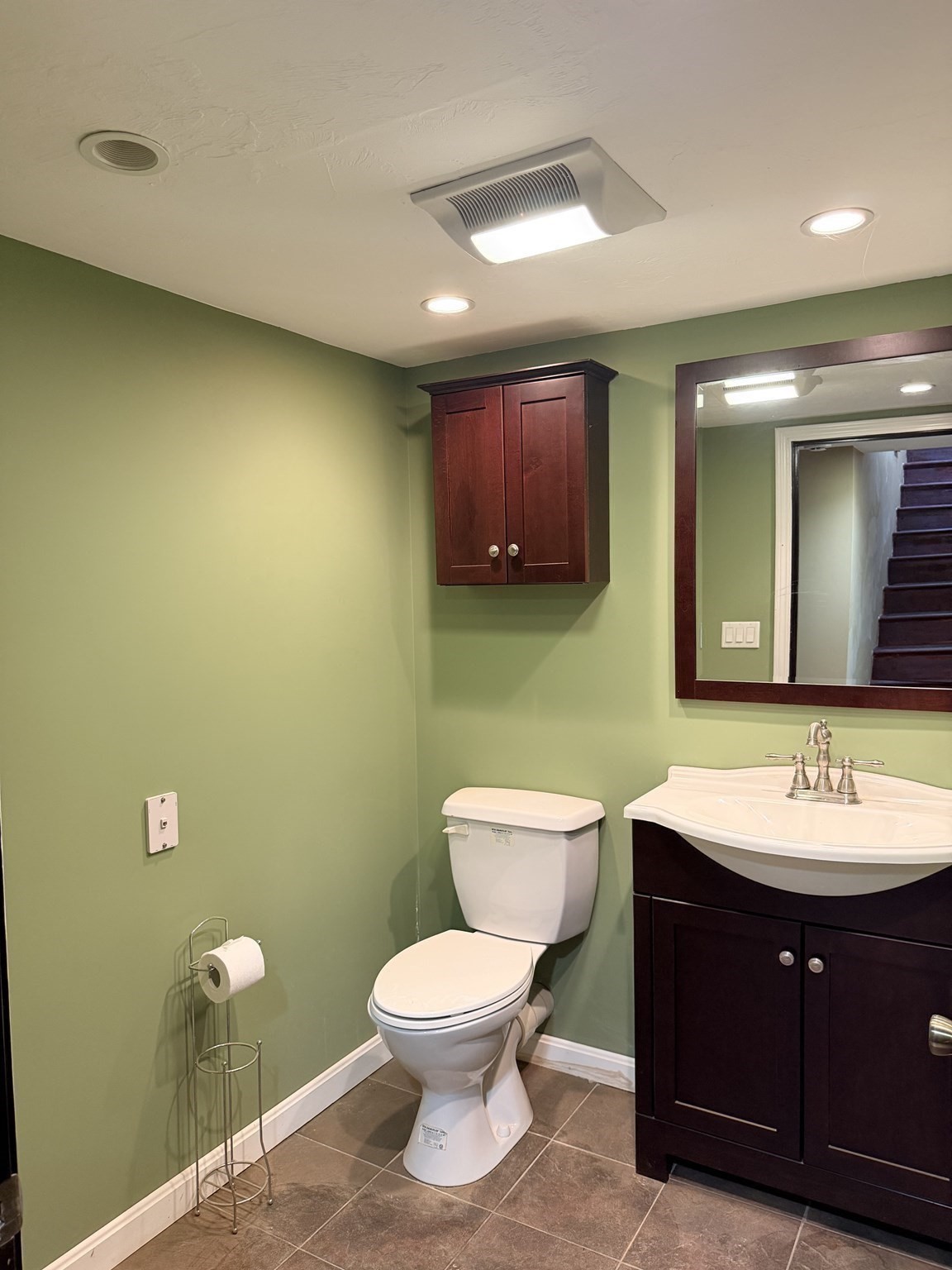 Bathroom with sage-green walls, dark wood vanity and cabinet, white toilet, and brown tile floor.