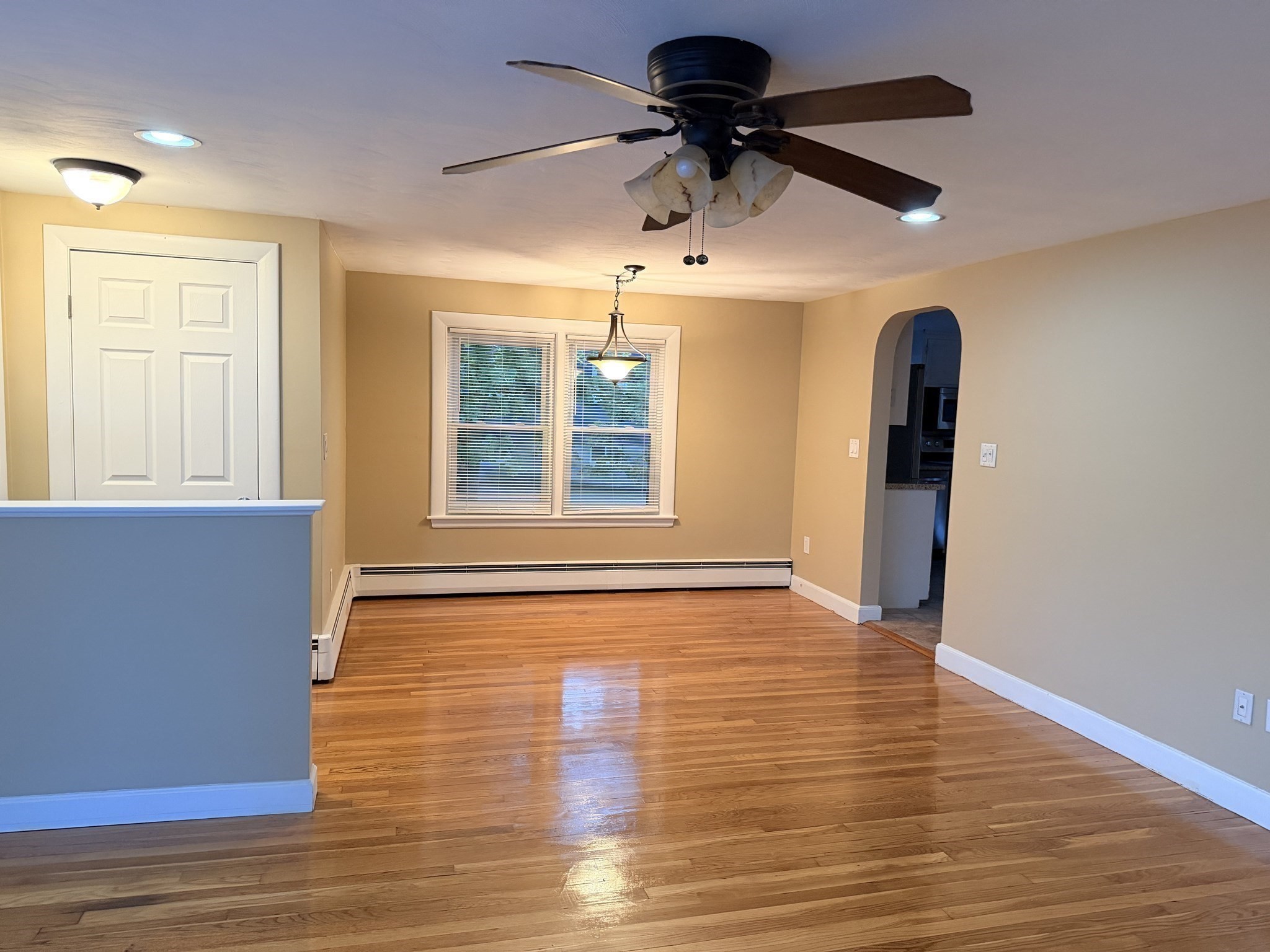 Empty dining area with polished hardwood floors, a window with blinds, and an arched kitchen opening.