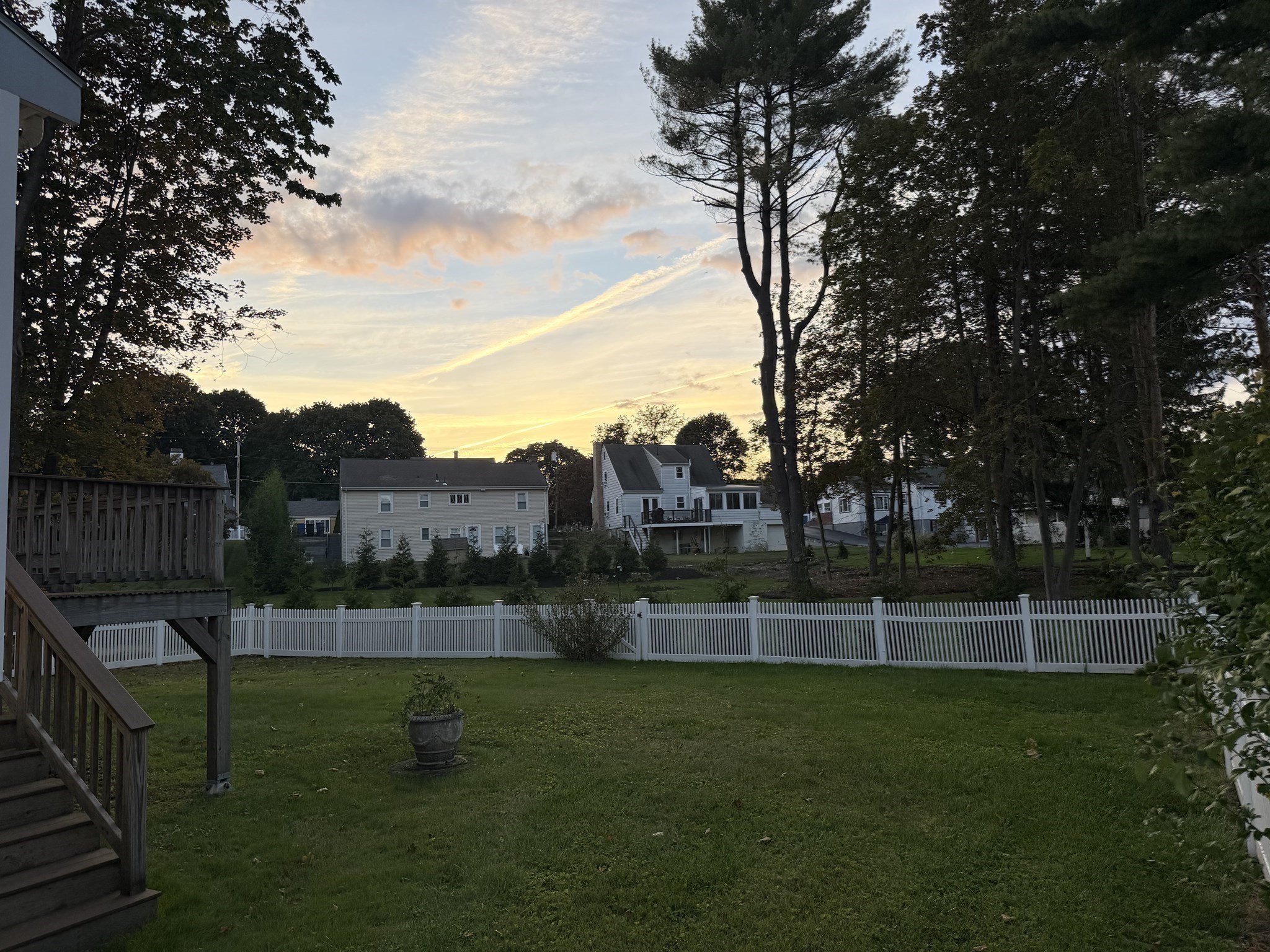 Backyard lawn and white fence beneath a sunset sky, viewed from a wooden deck.