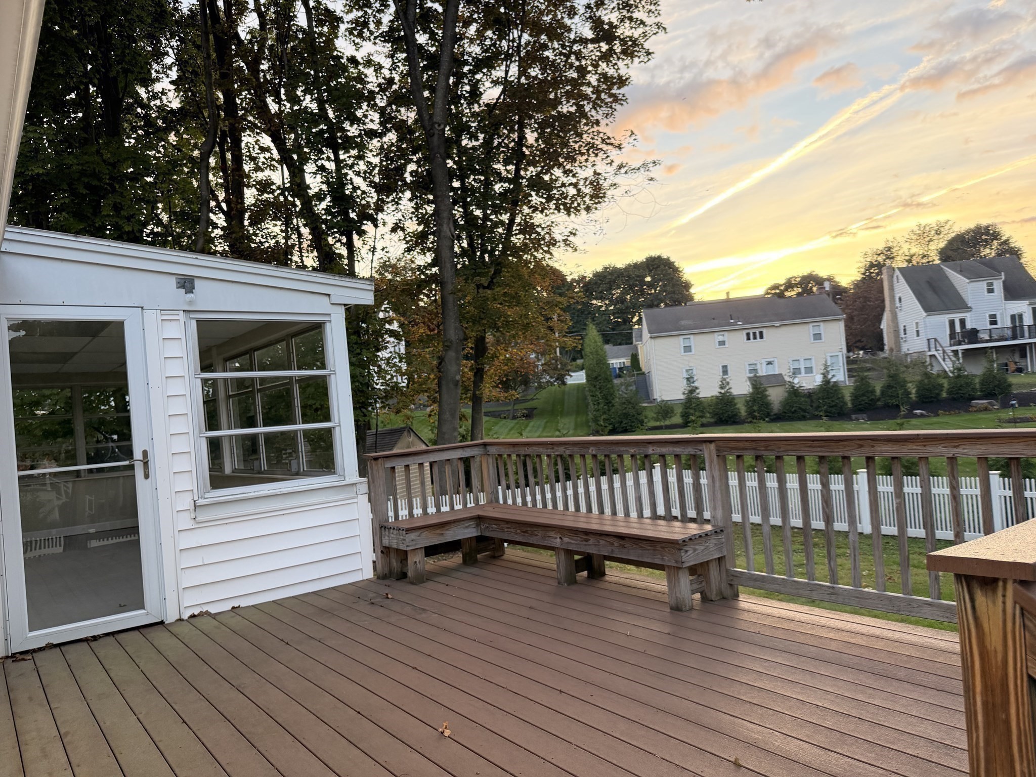 Spacious wooden deck with a built-in bench overlooking a lawn and neighboring homes at sunset.