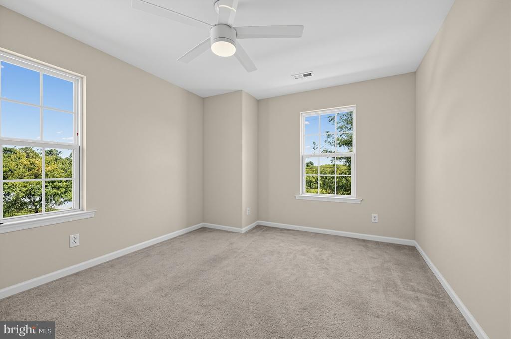 Empty bedroom with beige walls, carpet, two windows, and a ceiling fan.