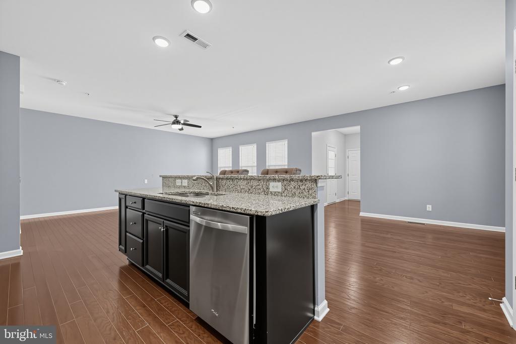 Open kitchen with a granite island, dark cabinets, stainless steel dishwasher, and brown wood-look flooring.