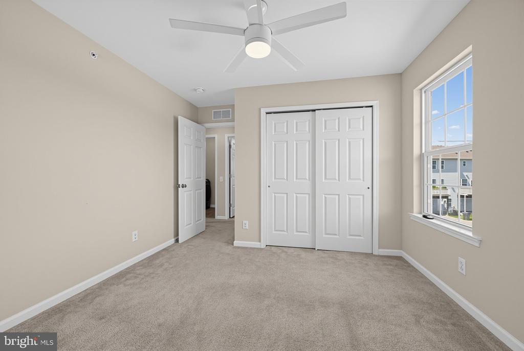 Empty carpeted bedroom with double closet doors, a ceiling fan, and a large window.
