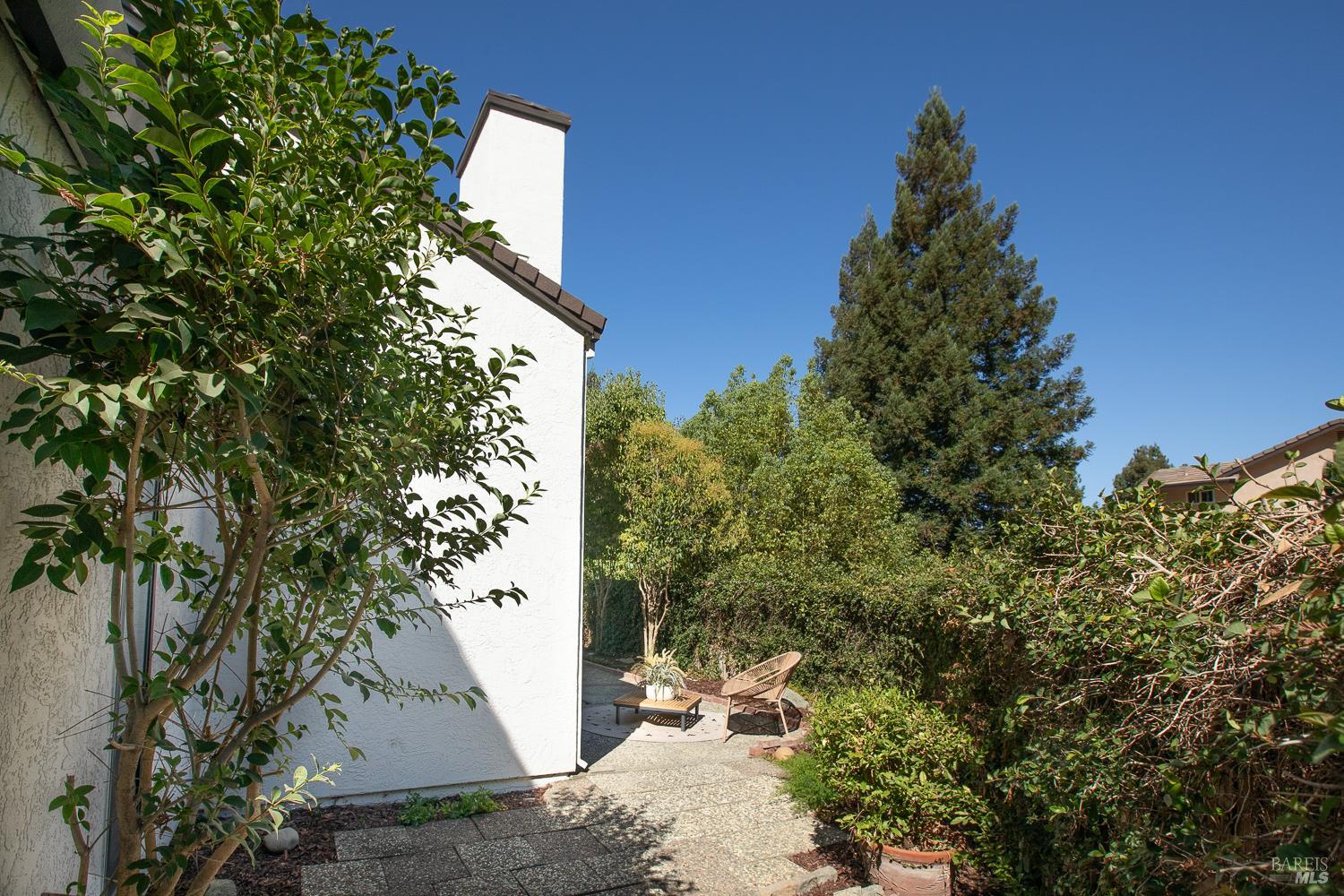 Side patio with outdoor seating beside a white stucco home, surrounded by mature greenery.