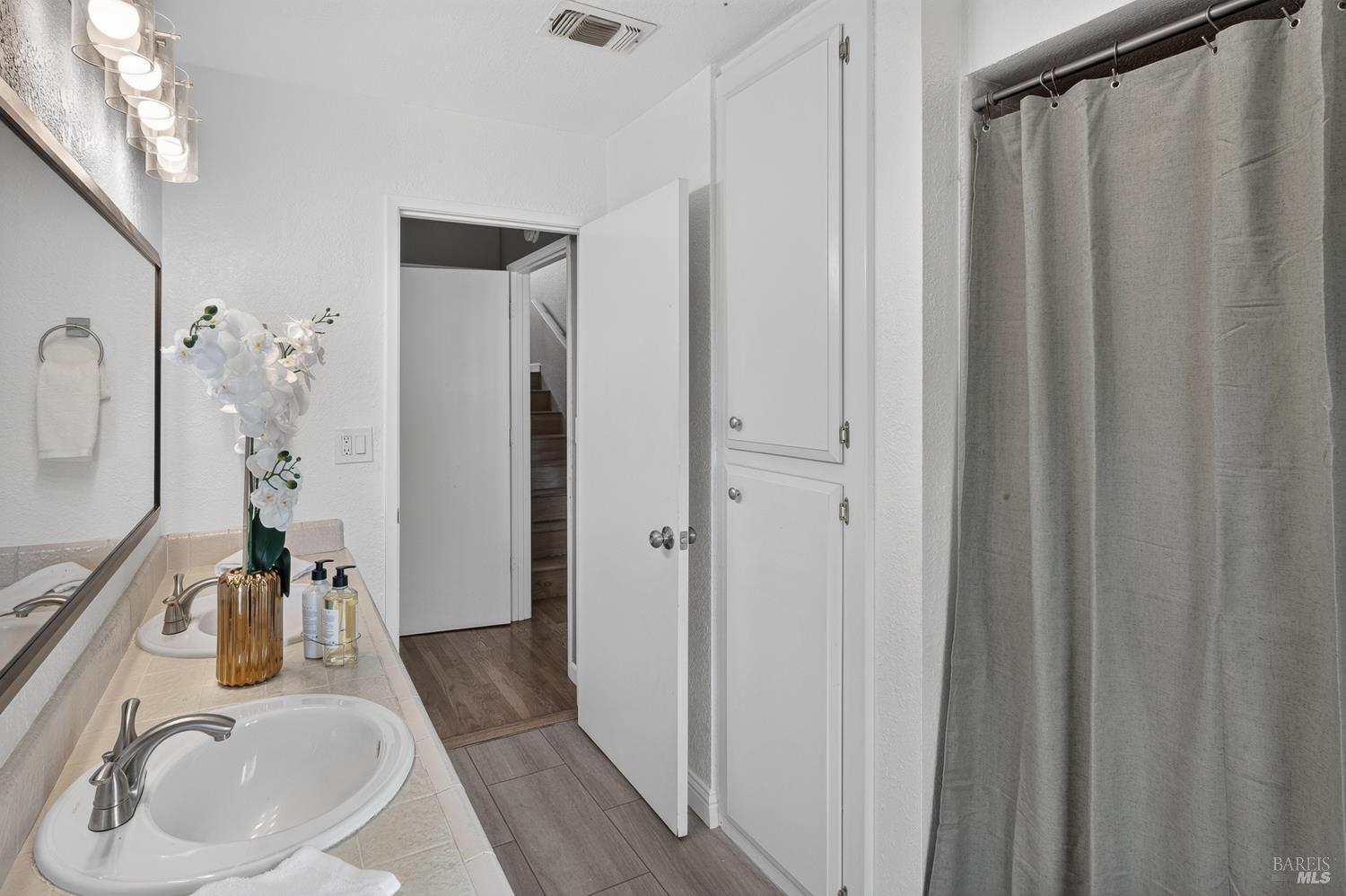 Bathroom with a two-sink tiled vanity, large mirror, gray shower curtain, and built-in linen cabinets.
