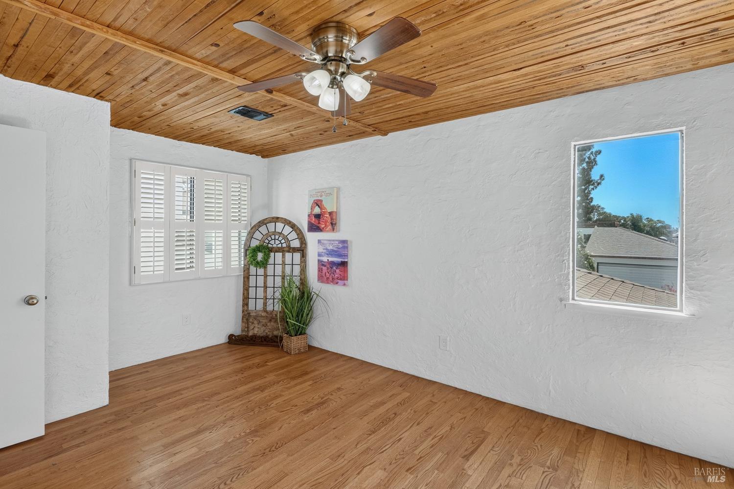 Empty room with hardwood floors, a wood-plank ceiling, ceiling fan, and white plantation shutters.