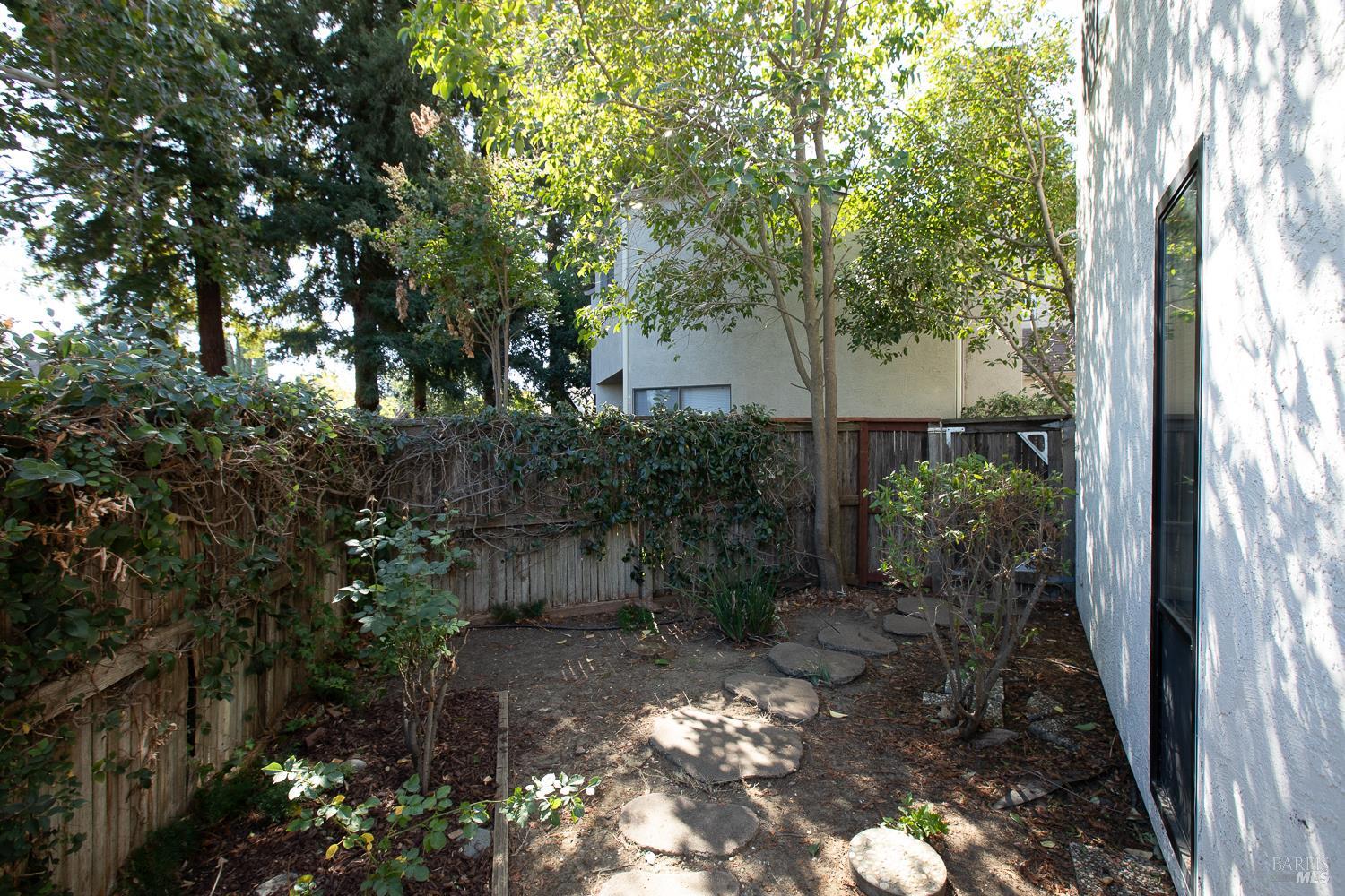 Side yard with round stepping stones, leafy shrubs, mature trees, and a wooden fence beside the house.