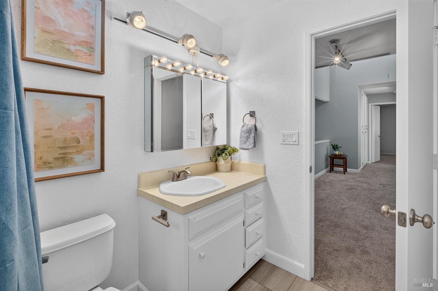 Bathroom with a beige-tiled vanity, mirrored medicine cabinet, toilet, and blue shower curtain.