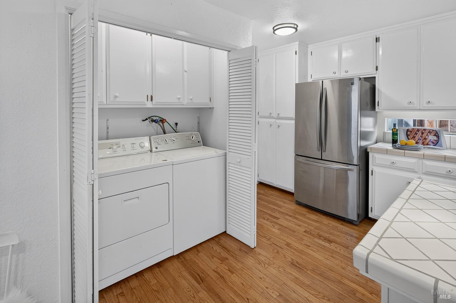 Kitchen with washer and dryer behind louvered doors, stainless steel refrigerator, and wood flooring.