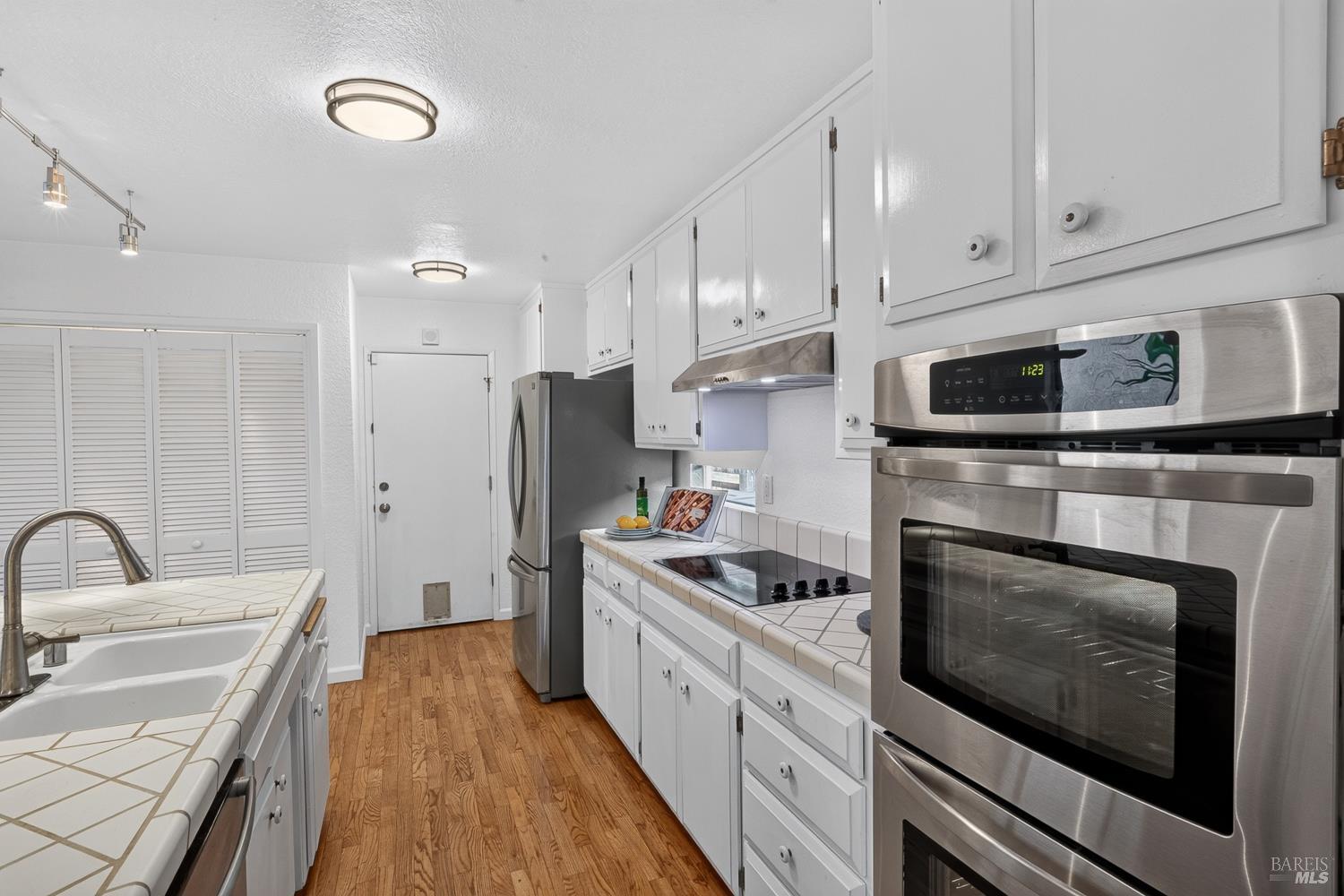 Galley kitchen with white cabinetry, stainless steel appliances, tiled counters, and hardwood flooring.