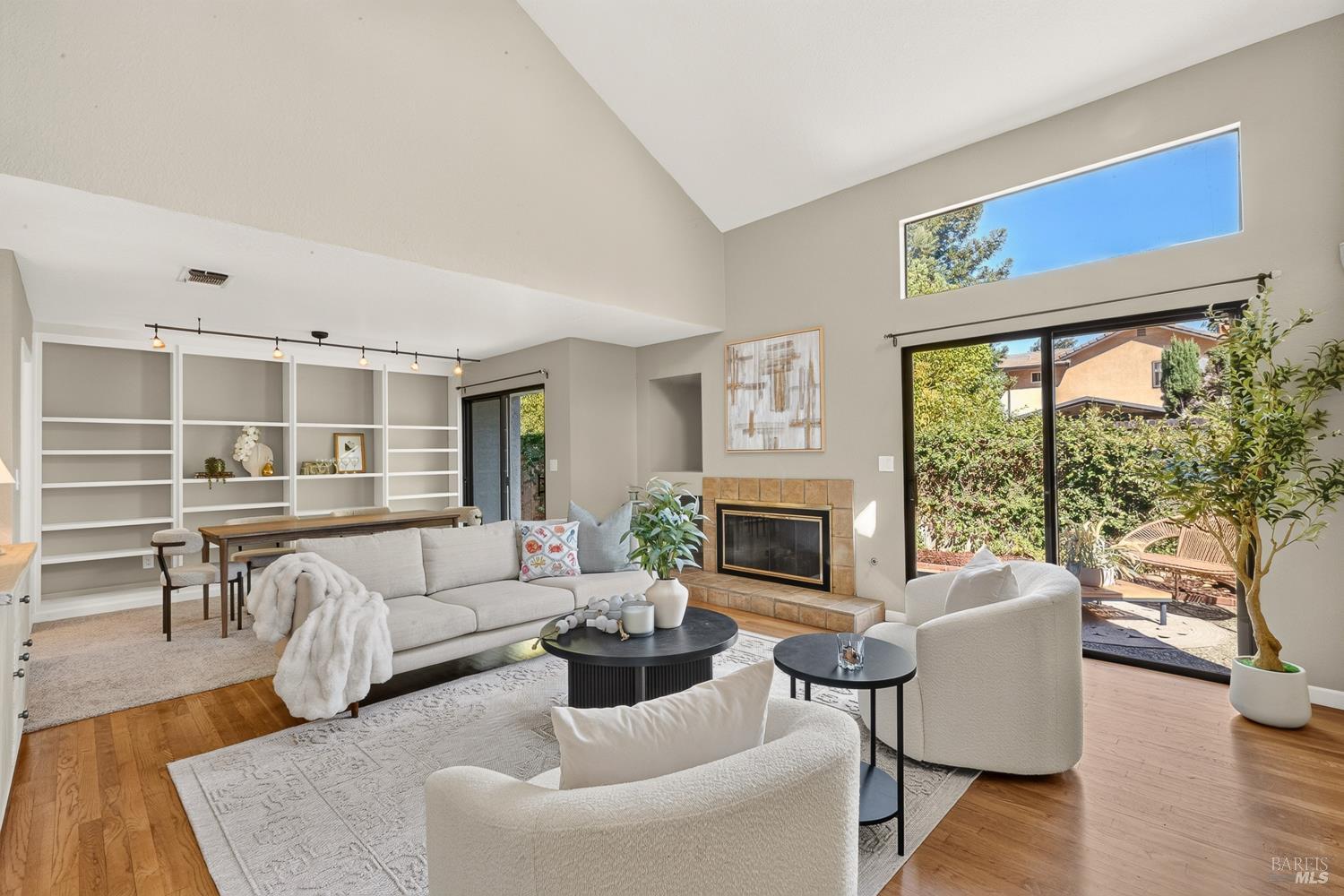 Living room with vaulted ceiling, tiled fireplace, built-in shelves, and garden-facing sliding glass doors.