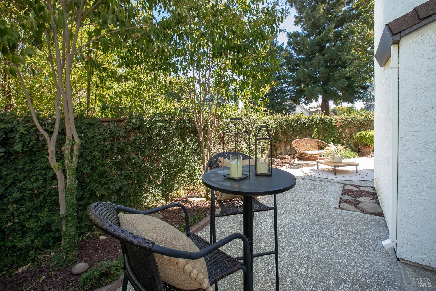 Backyard patio with a round black table, wicker chairs, mature trees, and a tall hedge.