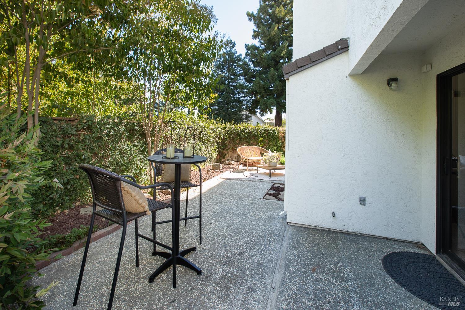 Patio beside a white stucco home with bistro seating and a leafy privacy hedge.