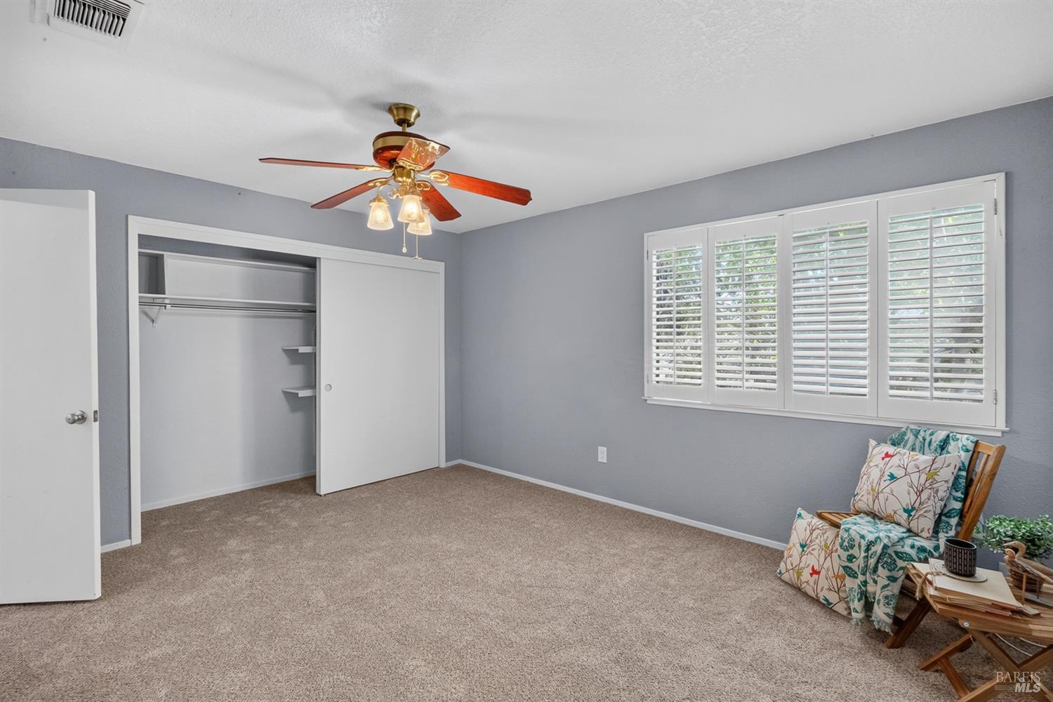 Bedroom with beige carpet, an open sliding-door closet, white plantation shutters, and a ceiling fan.