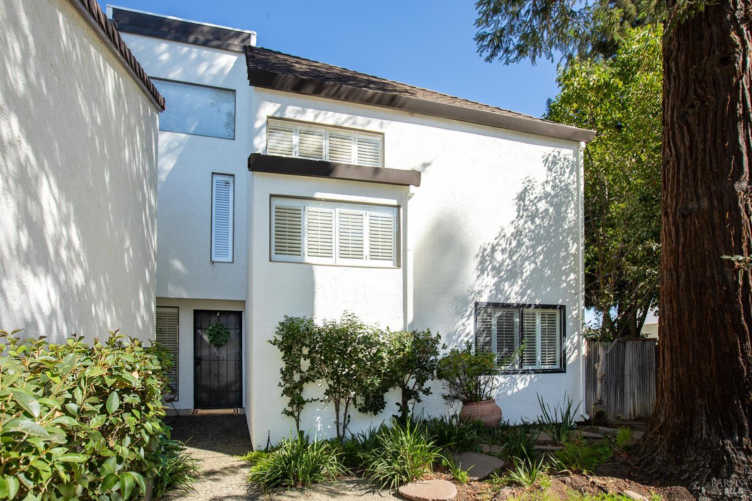 Two-story white stucco home with shuttered windows, garden beds, and a large tree beside it.