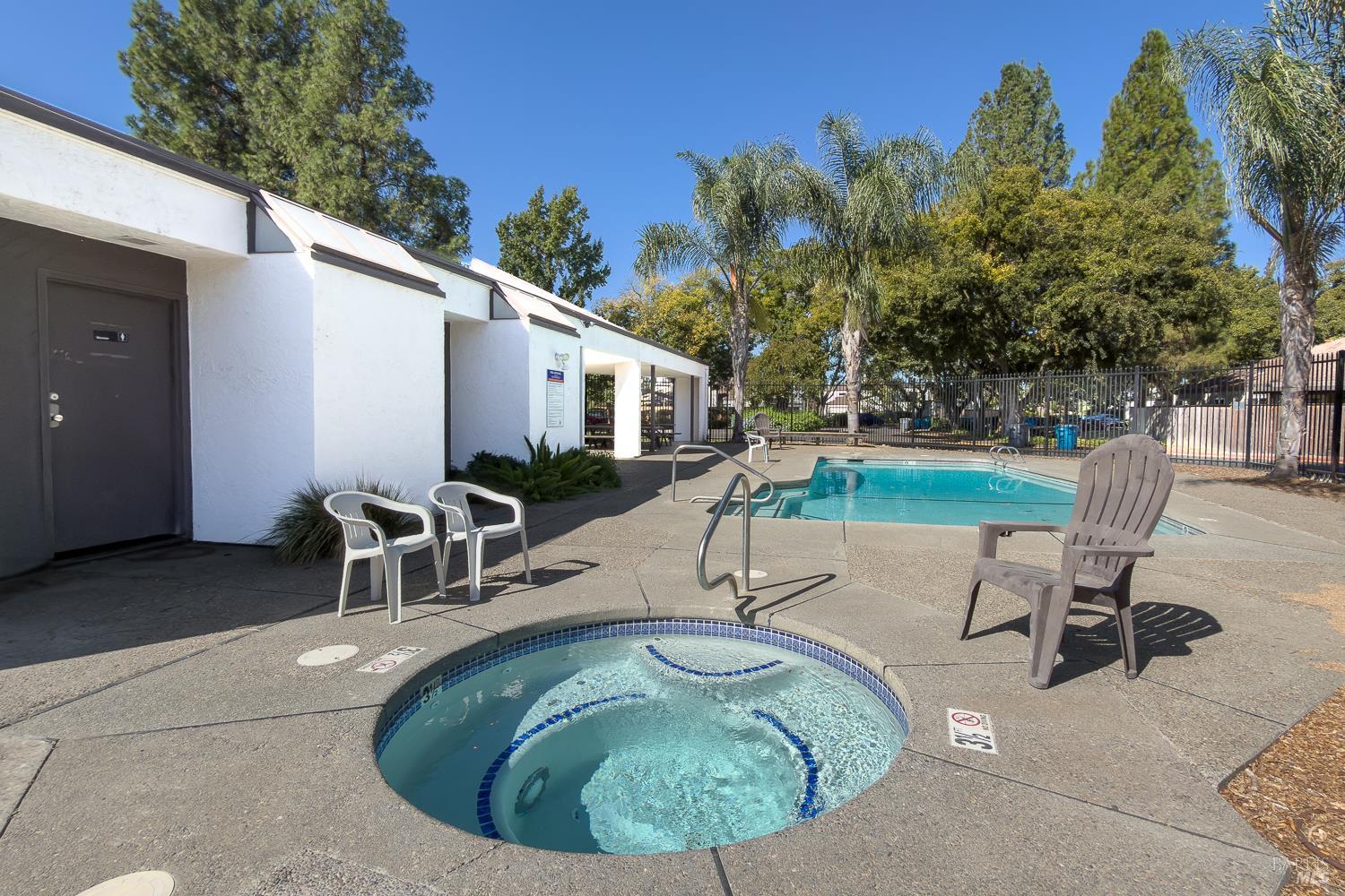 Outdoor pool and circular spa beside a white building, with a fenced deck and palm trees.