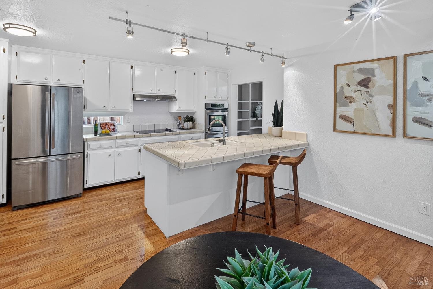 Kitchen with white cabinets, stainless appliances, a tiled island, wooden stools, and hardwood floors.