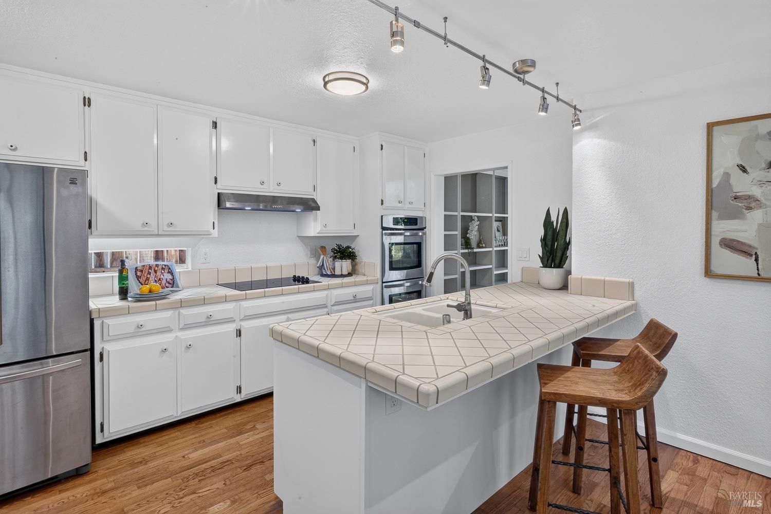 Kitchen with a large tiled island, white cabinetry, stainless appliances, and wooden bar stools.