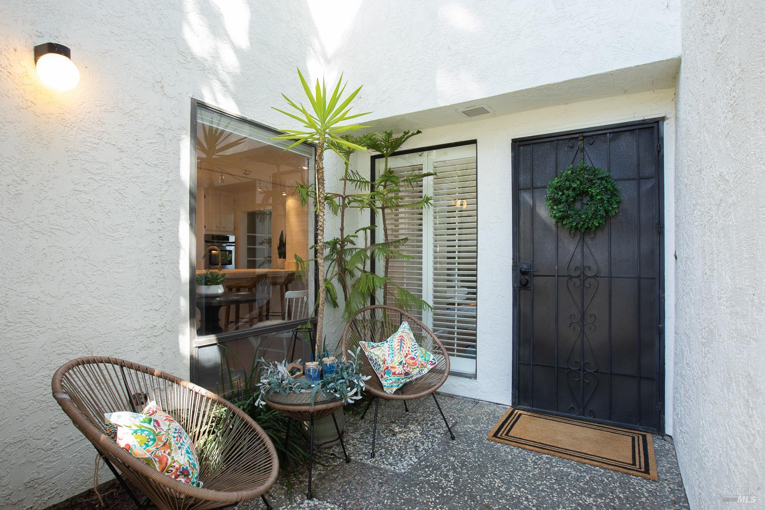 Home entry patio with a black metal door, tropical plants, and colorful cushions on wicker chairs.