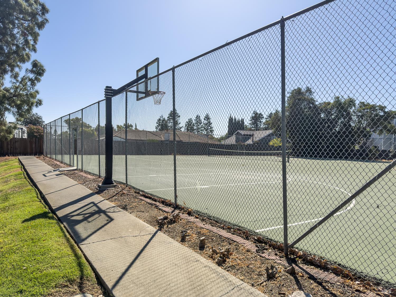 Fenced outdoor court with a basketball hoop and painted lines beside a concrete walkway.