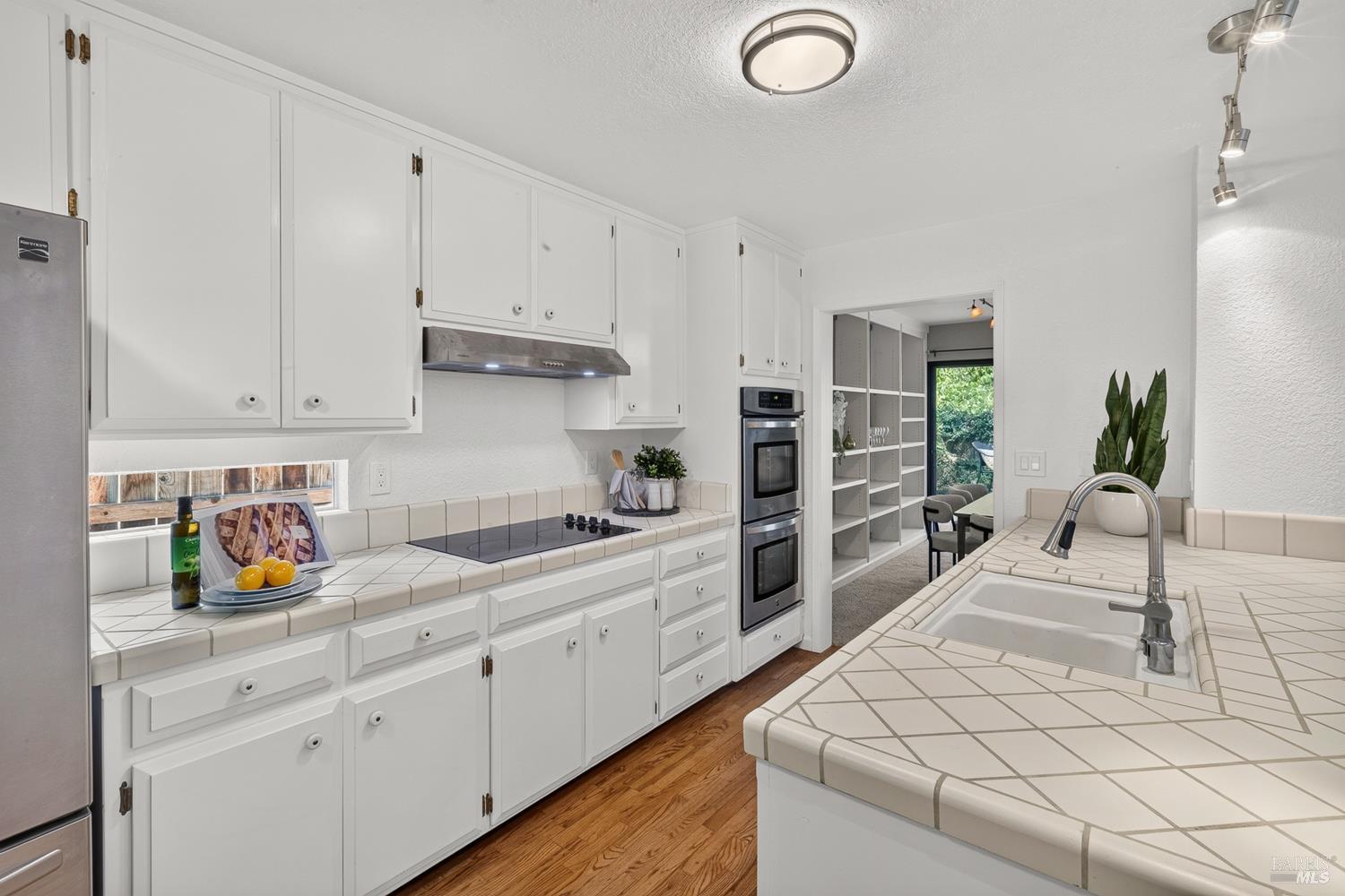 Bright kitchen with white cabinetry, tiled island and counters, stainless steel double ovens, and hardwood flooring.