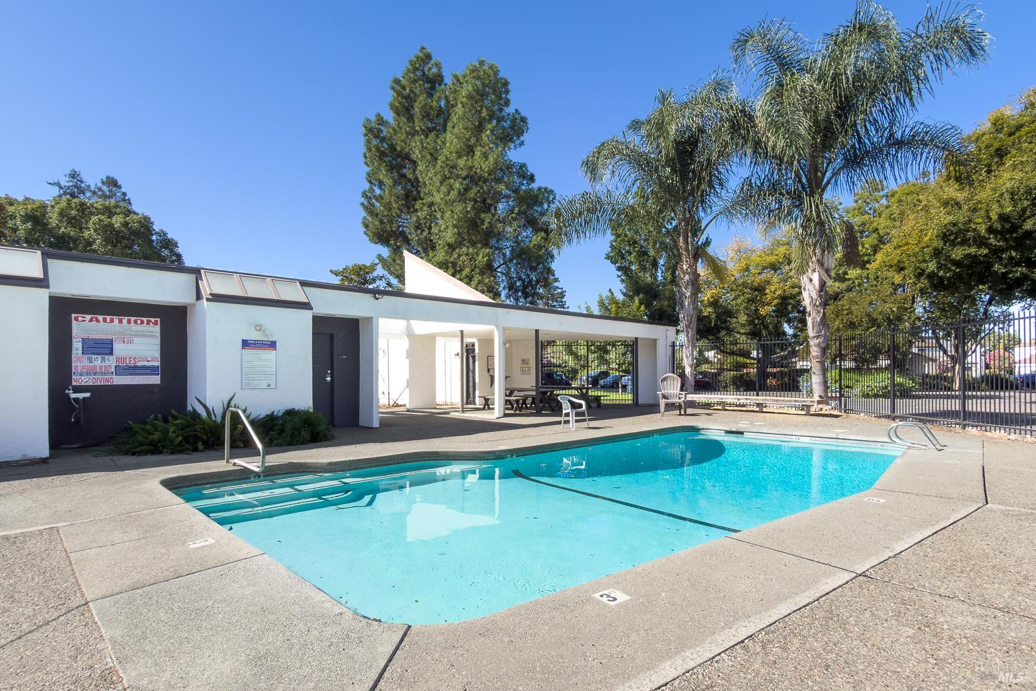 Outdoor swimming pool beside a white pool building, with palm trees and a fenced deck.
