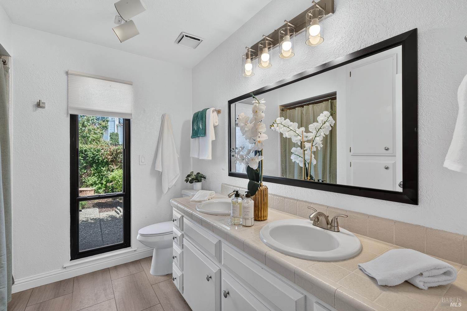 Bathroom with a double-sink tiled vanity, large black-framed mirror, and window overlooking greenery.