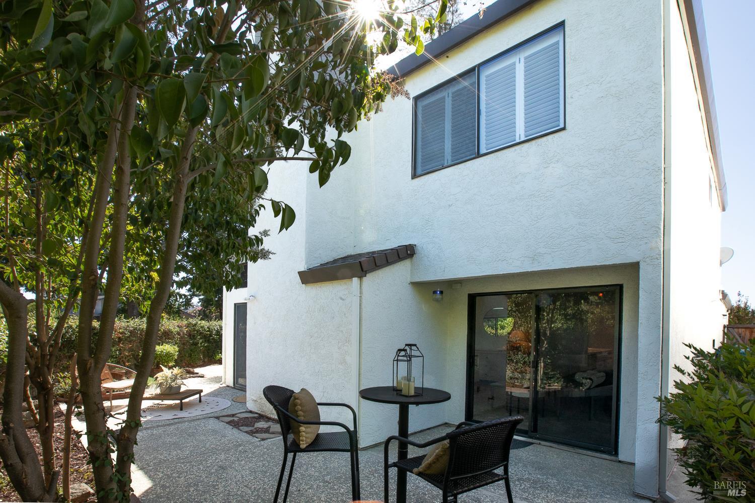 Two-story stucco home with a shaded patio, sliding glass door, and outdoor seating beneath mature trees.