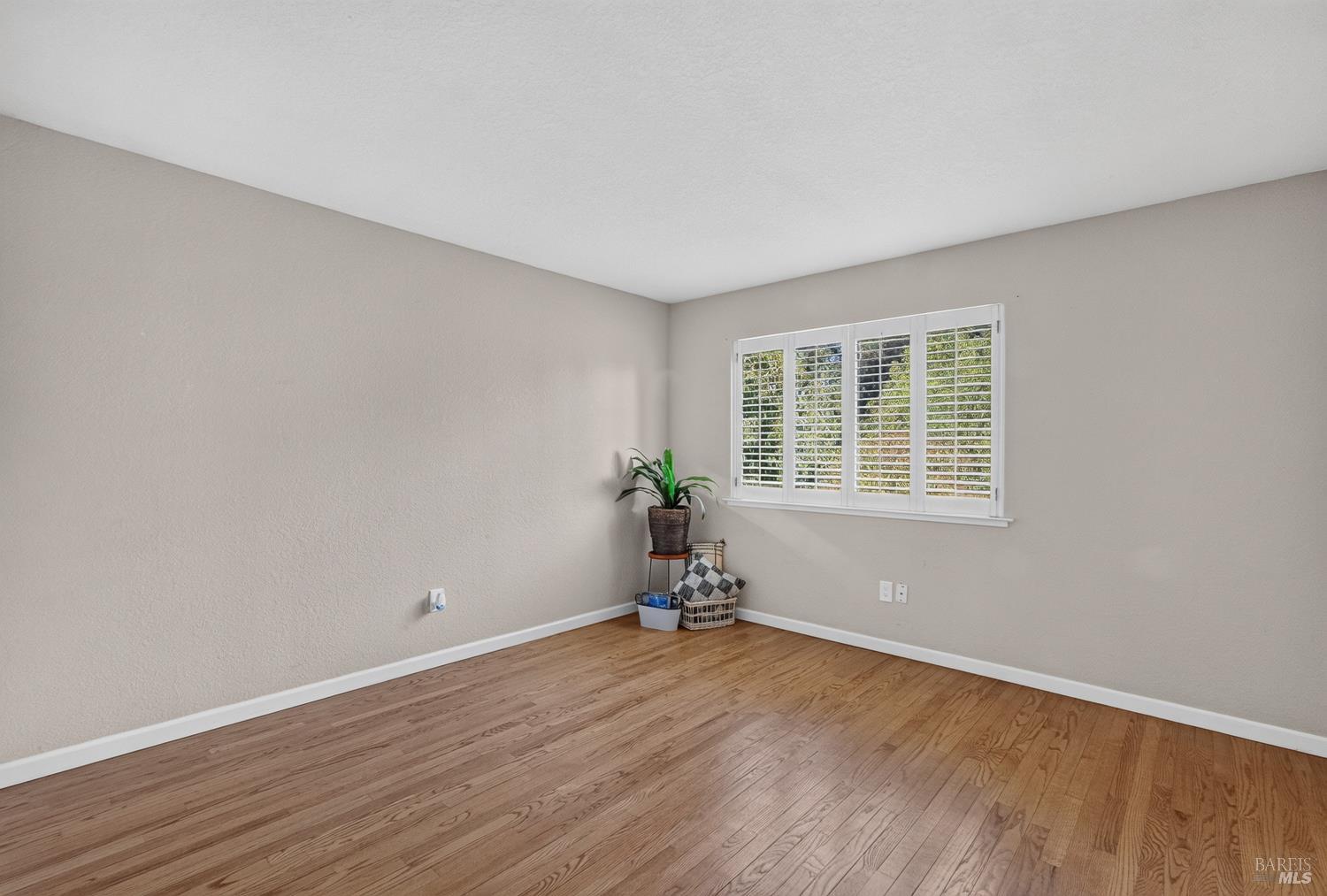 Mostly vacant room with hardwood floors and a wide window with white plantation shutters.
