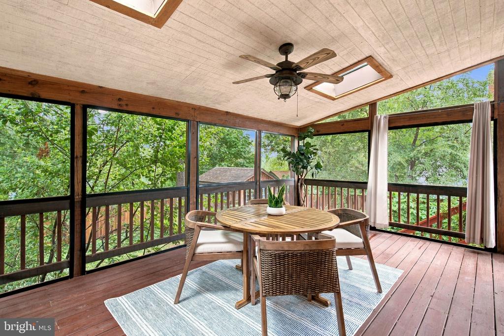 Screened-in porch with a dining set, ceiling fan, skylights, and leafy wooded views.