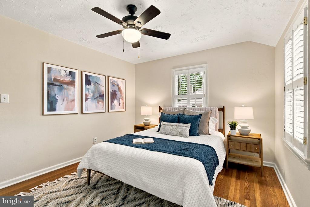 Staged bedroom with a navy-and-white bed, abstract wall art, shuttered windows, and hardwood floors.