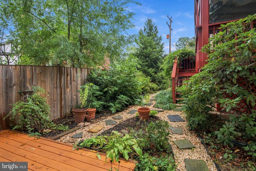 Landscaped backyard with a wooden deck, gravel stepping-stone path, and dense greenery beside a red-sided home.