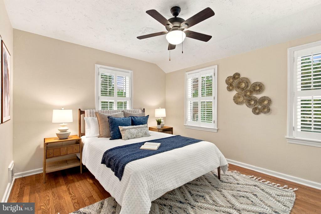 Staged bedroom with a white bed, navy accents, shuttered windows, and hardwood floors.