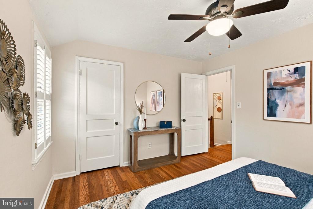 Staged bedroom with hardwood floors, plantation shutters, and a blue-covered bed beneath a ceiling fan.