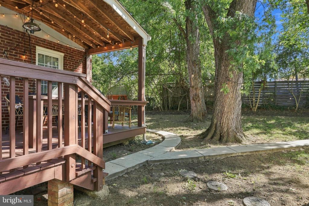 Covered wooden deck with string lights beside a shaded backyard path winding around mature trees.