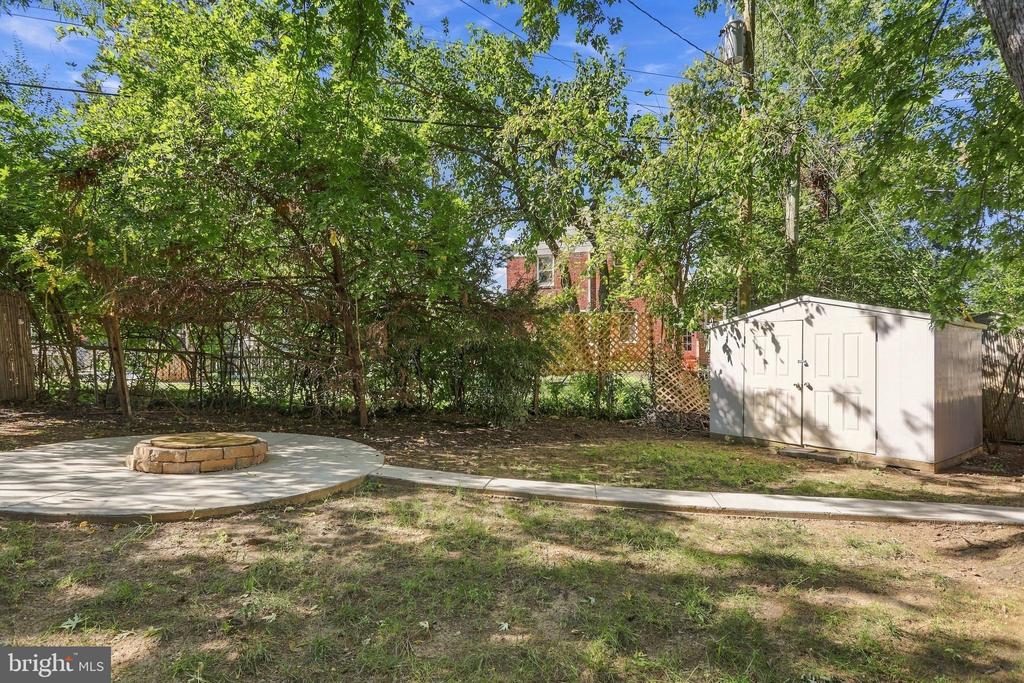 Backyard patio with a circular stone fire pit, white storage shed, and mature trees.