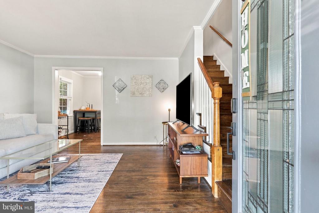 Living room with a light sofa, hardwood floors, and a staircase beside the open front door.