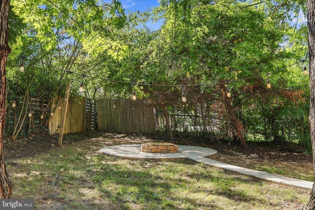 Fenced backyard patio with a circular stone fire pit, mature trees, and string lights.
