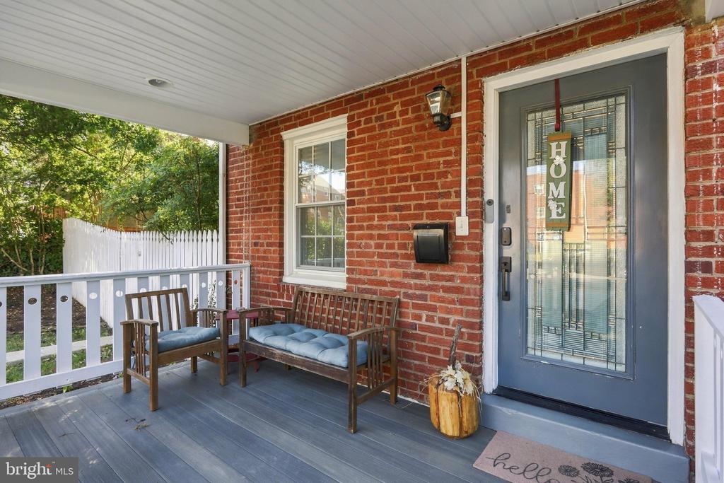 Covered brick front porch with a blue glass-paneled door, white railing, and wooden seating.