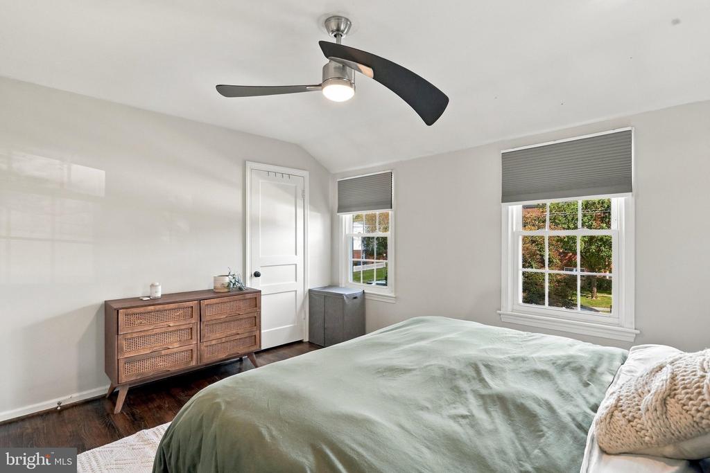 Bedroom with pale green bedding, a wooden dresser, and two windows overlooking trees.
